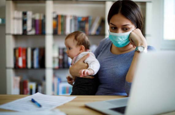 A woman wearing a disposable face mask holds a baby while sitting at a desk.