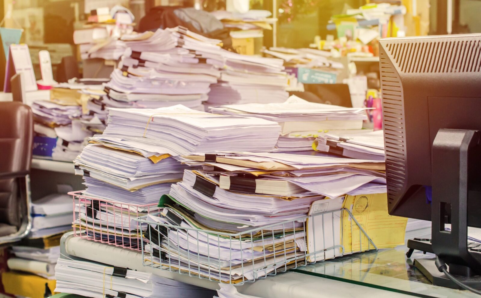 Many large stacks of paper cluttering the top of office desks.