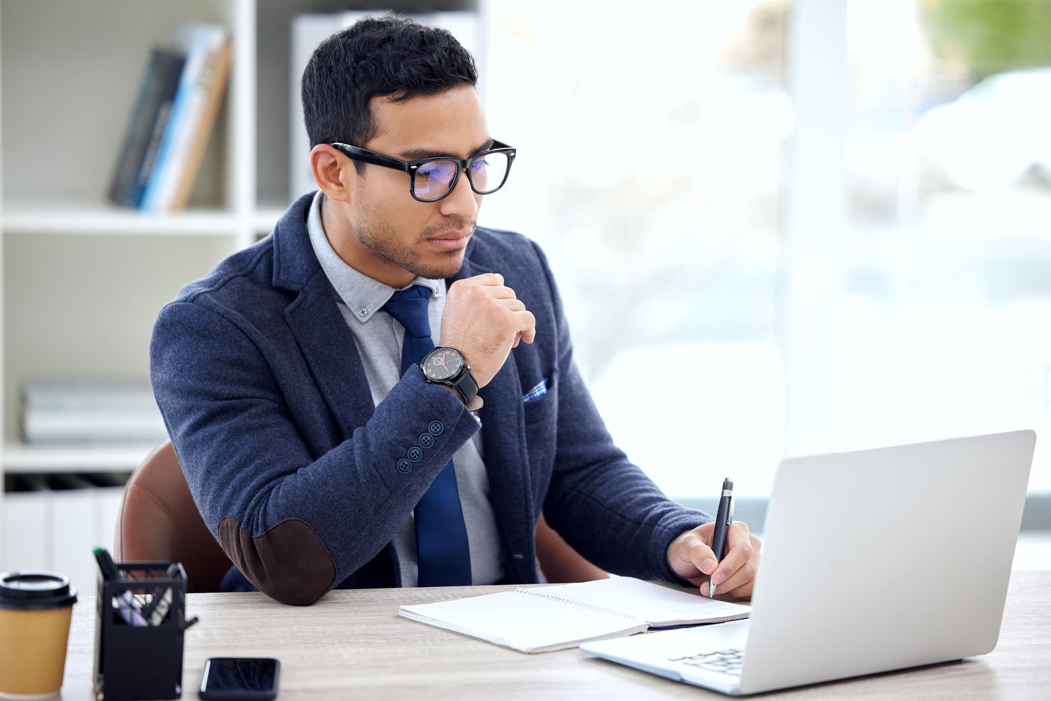 Man wearing suit with glasses staring at laptop taking notes by hand on a piece of paper